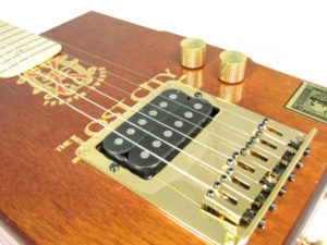 This electric cigar box guitar features a bottom-loading gold hard-tail bridge with humbucker opening. 